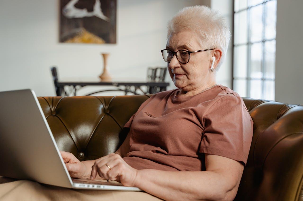 Senior adult woman with glasses using a laptop on a comfortable couch indoors.